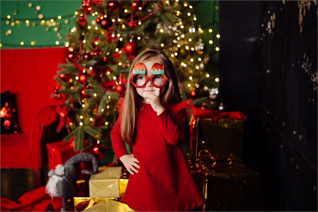 une petite fille avec une robe rouge et des lunettes rouge, se tient devant un sapin de Noël - freep