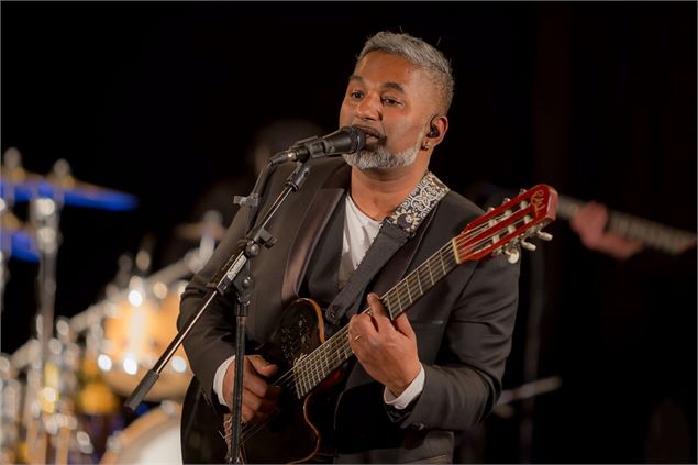 Un homme en costume noir chante et joue de la guitare acoustique sur scène. Une batterie est visible