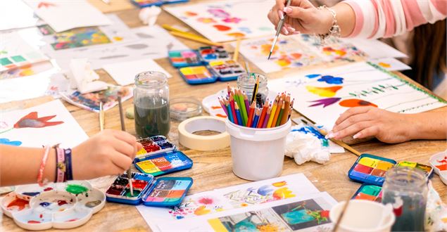 Zoom sur une table recouverte d'ustencils créatifs, mains d'enfant et d'adulte en train de peindre -