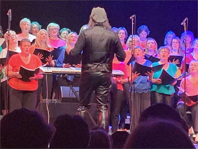 Un groupe de personnes chante, debout en rangs, visage éclairés par des lumières colorées. Au premie