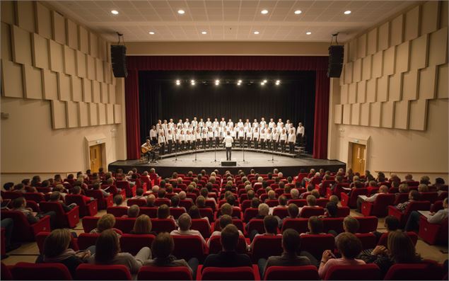 Vue depuis le public d'un auditorium moderne où une chorale en blanc chante sur scène face à une sal