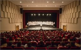 Vue depuis le public d'un auditorium moderne où une chorale en blanc chante sur scène face à une sal
