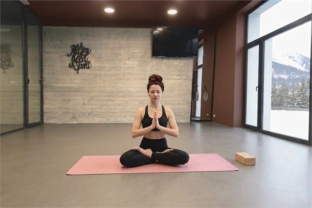 Une femme brune en méditation, mains jointes, assise au centre d'une salle de yoga épurée face à des