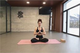 Une femme brune en méditation, mains jointes, assise au centre d'une salle de yoga épurée face à des