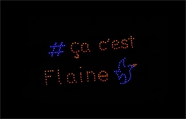 Spectacle nocturne de drones sur fond noir. Des points lumineux forment le texte orange « # ça c’est