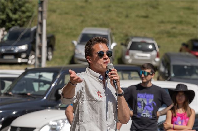 Un commentateur avec un micro et lunettes de soleil devant des spectateurs et un parking - Flo Littl
