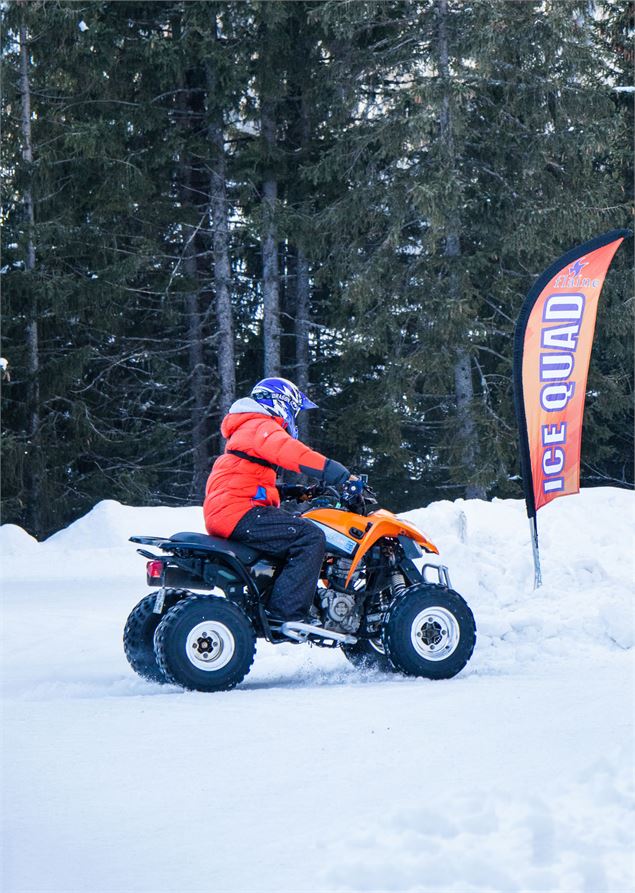 Une personne en veste orange et casque bleu conduit un quad noir et orange de profil. Le véhicule ro