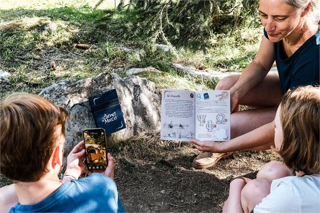 Un adulte et deux enfants sont assis par terre en forêt et lisent ensemble le carnet de Marcel - OT 